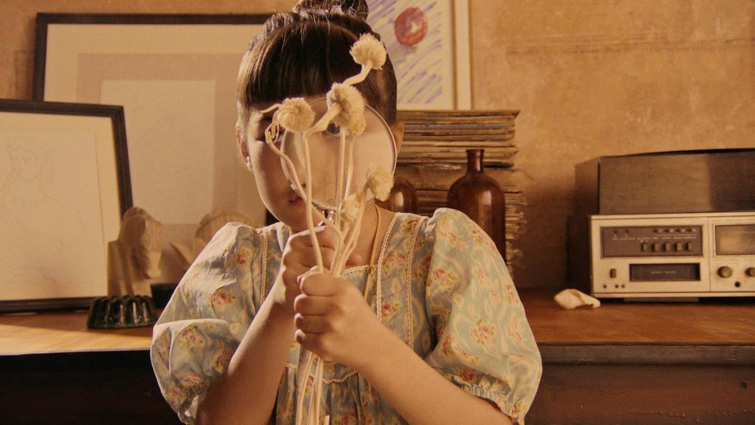 A young girl in a floral dress is holding a bouquet of white flowers in front of her face in a warmly lit, vintage-style indoor setting.