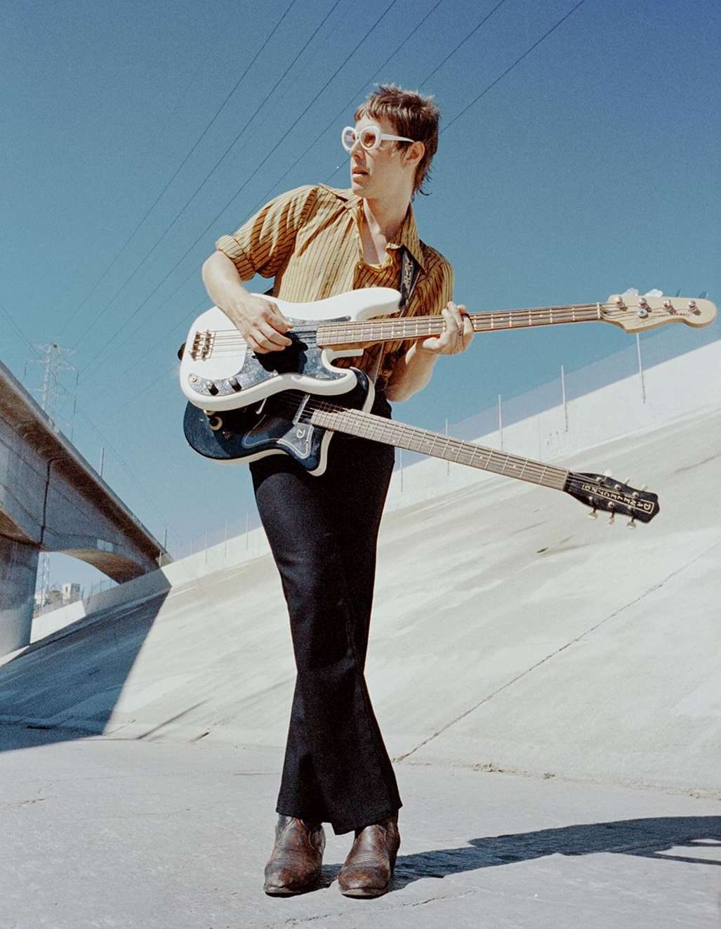 Adult male musician stands outdoors playing a double-neck bass guitar wearing vintage sunglasses, brown boots, and a striped shirt.