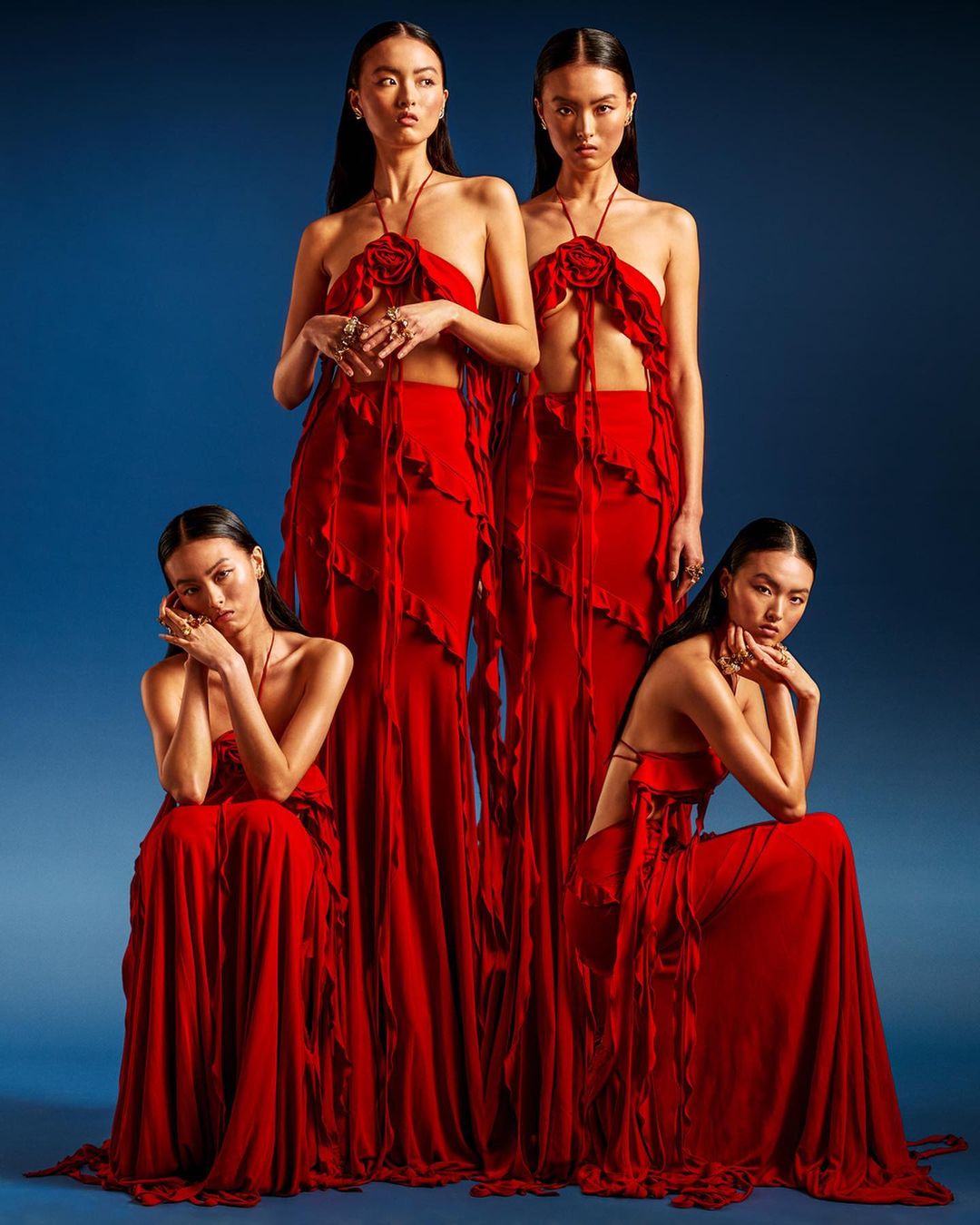 Group fashion portrait of four Asian women wearing long, red, rose-detailed dresses against a blue backdrop in a studio setting.