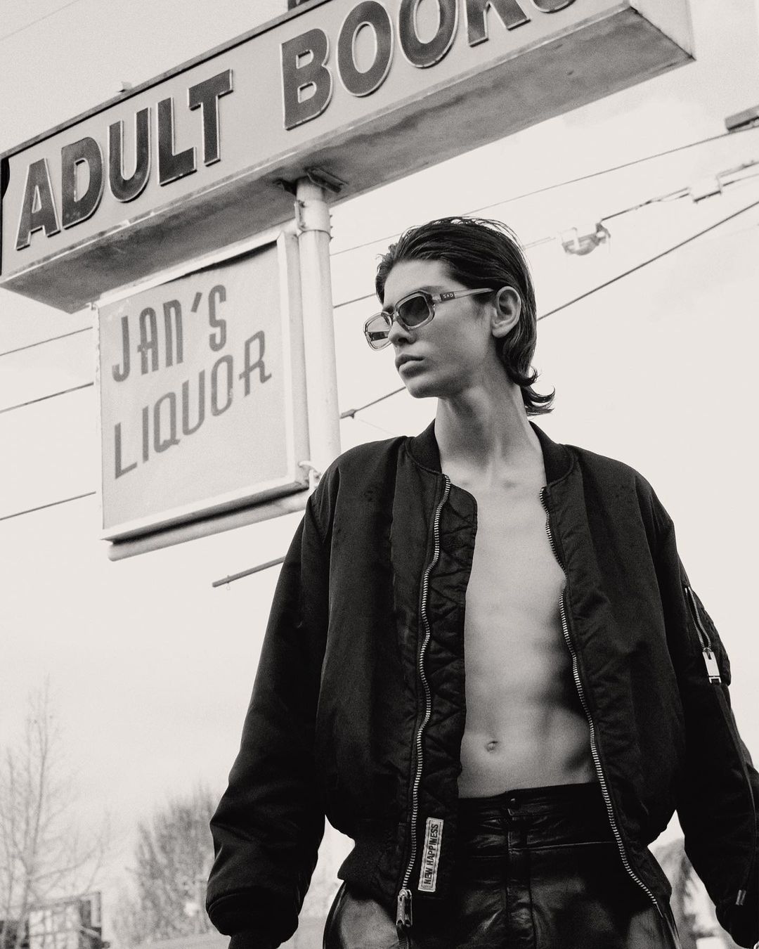 Monochrome portrait of a topless adult in a bomber jacket and sunglasses posing outdoors under an 'Adult Books' and 'Jan's Liquor' sign.