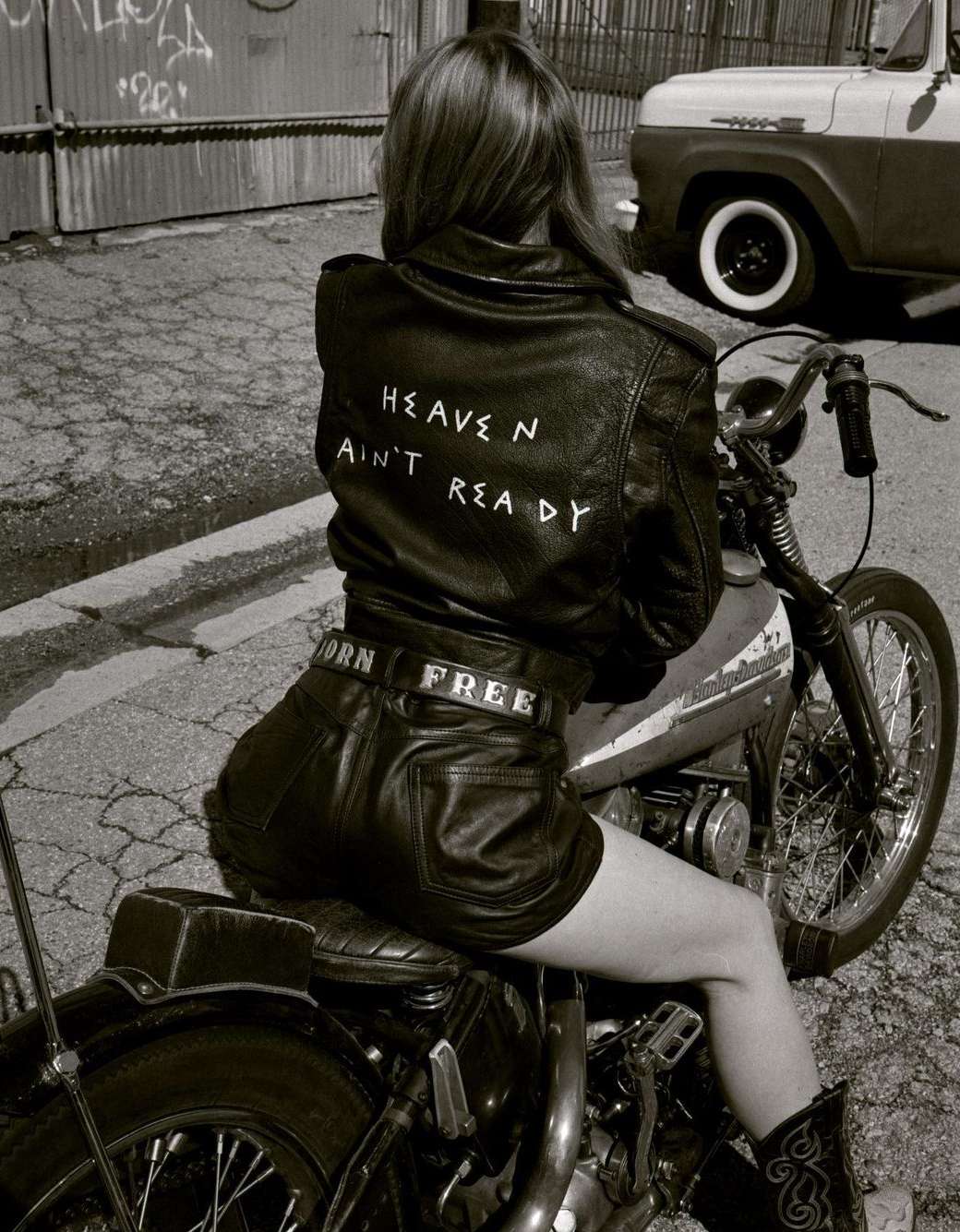 Back view of a woman in a leather jacket and shorts sitting on a motorcycle in an urban alleyway, with a vintage truck in the background.