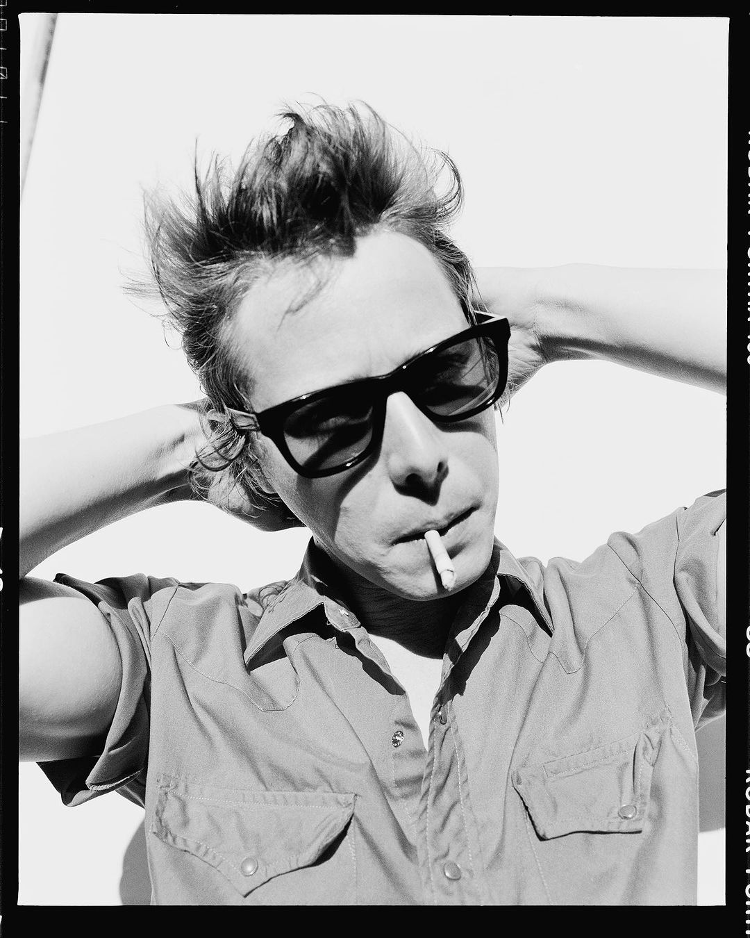 Monochrome portrait of a man with styled hair and sunglasses, cigarette in mouth, hands behind his head, wearing a collared shirt against a white wall.