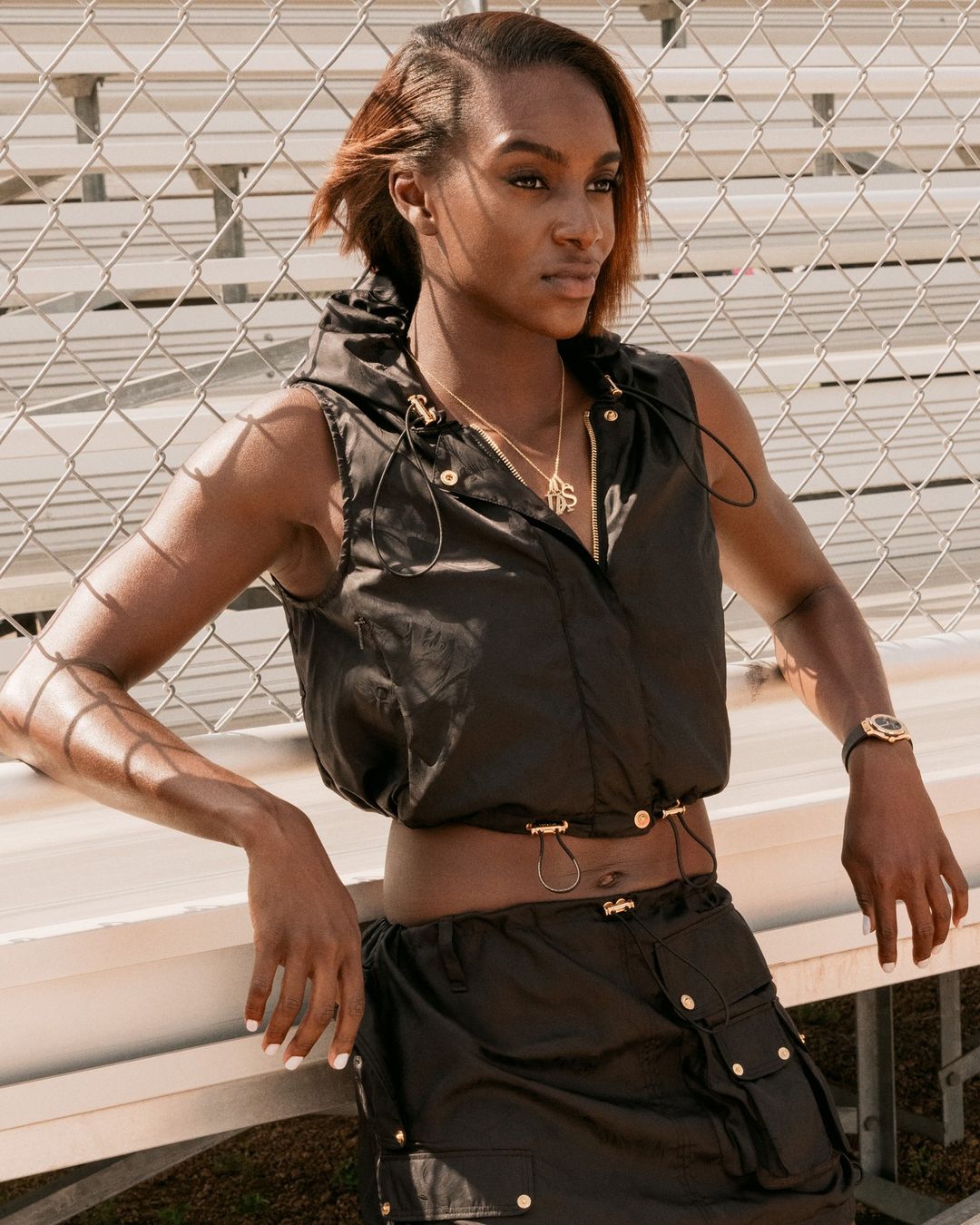 A slender, stylish woman poses confidently by a chain-link fence, showcasing a black, hooded vest and skirt set, accented by gold jewelry.