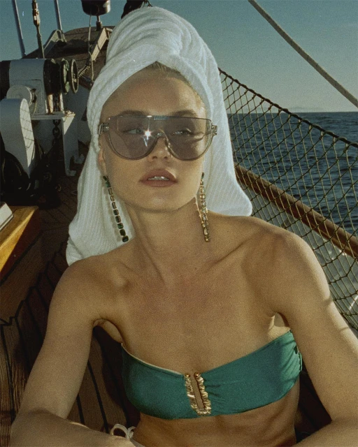 Woman in a strapless top and a towel on her head, wearing sunglasses and earrings, on a boat with water and blue sky background.