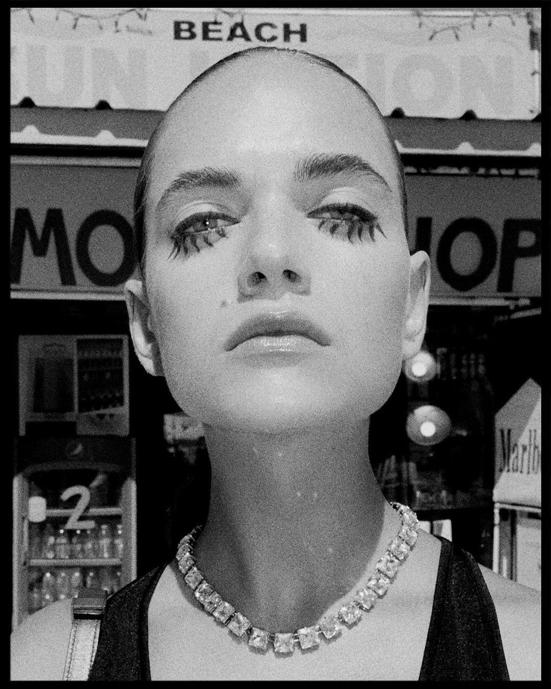 Striking black and white portrait of a bald adult female with bold makeup, a diamond necklace, and a serious facial expression.