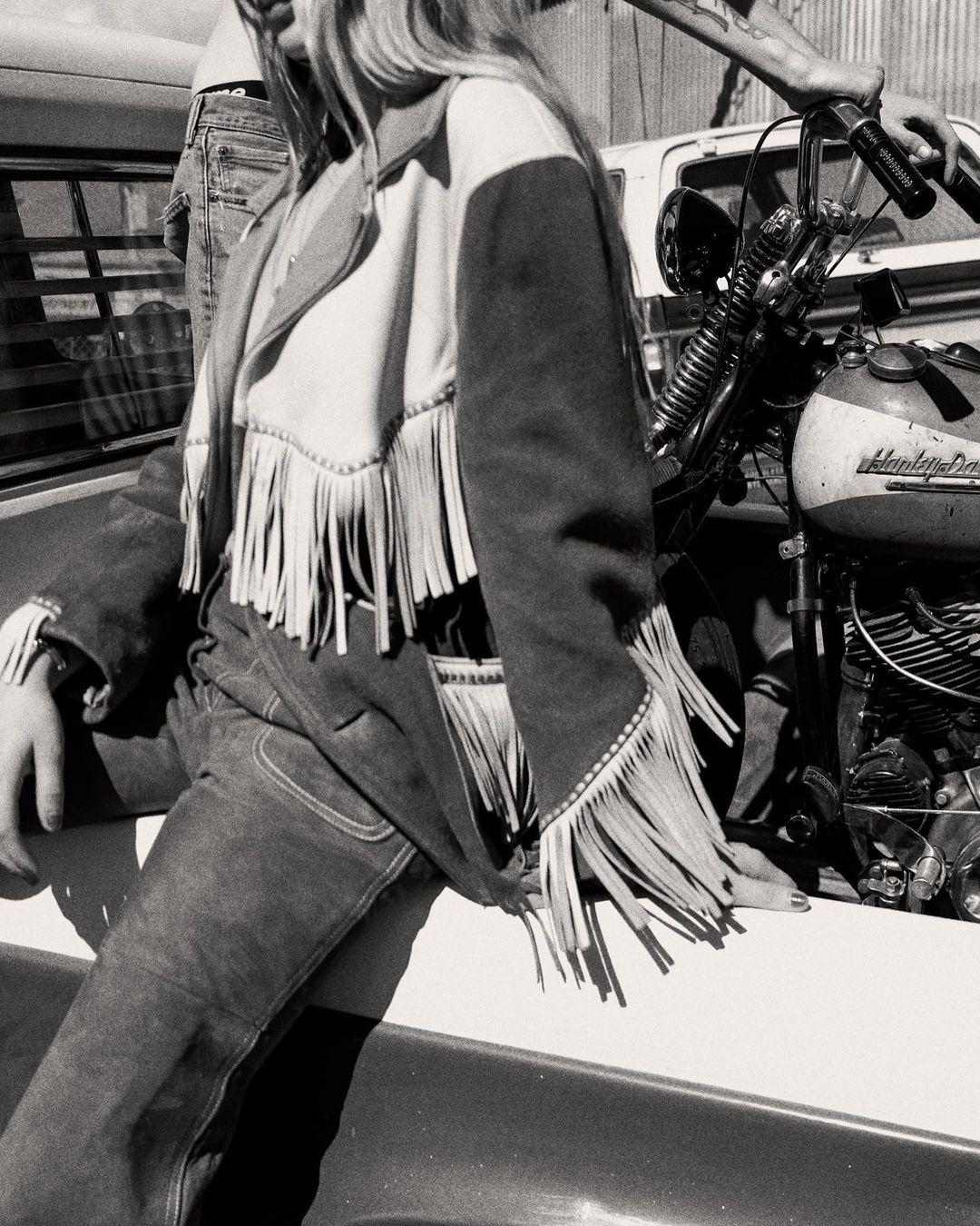 Black and white image featuring a woman wearing a fringed jacket and pants, with a motorcycle partially visible in the background.