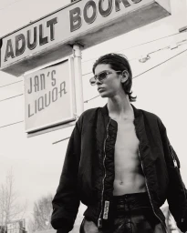 Monochrome portrait of a young man wearing sunglasses and an unzipped bomber jacket outdoors near signage.