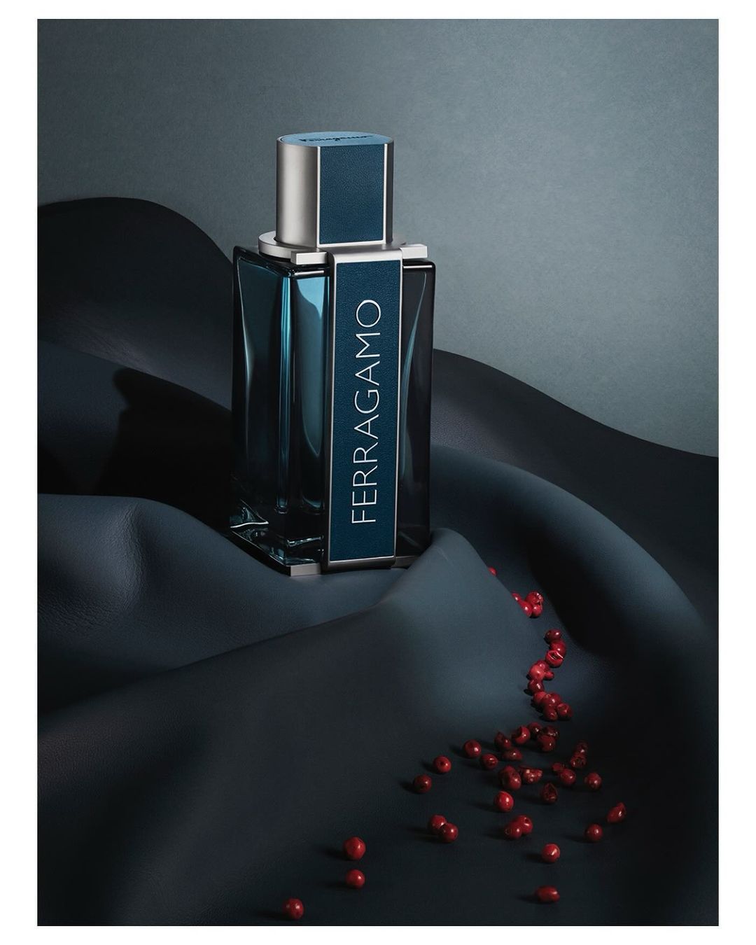 Ferragamo perfume bottle on rippled black fabric with scattered red berries, studio setting, grey background, product shot.