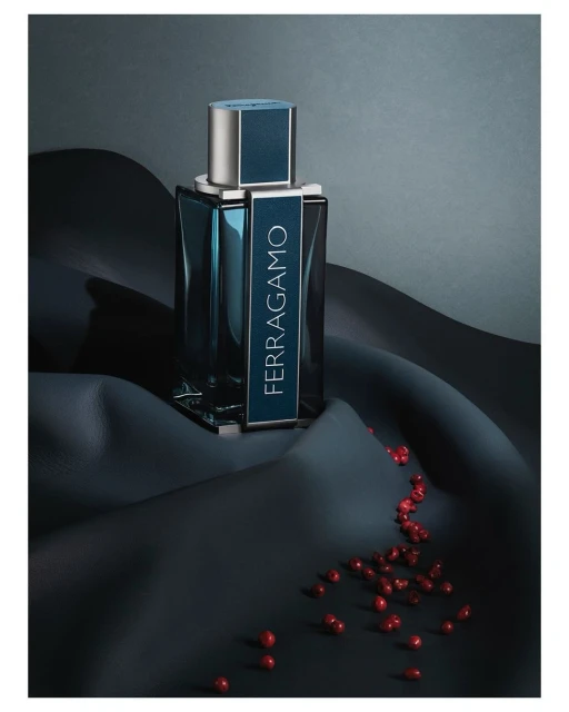 Ferragamo perfume bottle on rippled black fabric with scattered red berries, studio setting, grey background, product shot.