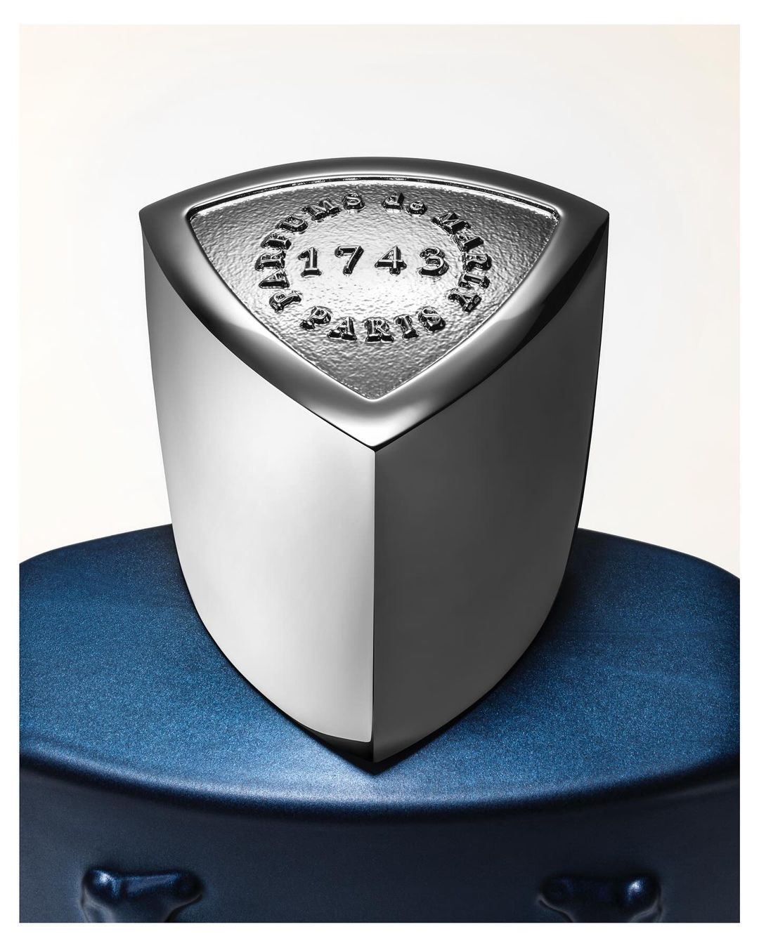 Close-up studio shot of a metallic and blue perfume bottle, featuring embossed text on a shield-shaped emblem. Luxury fragrance product.