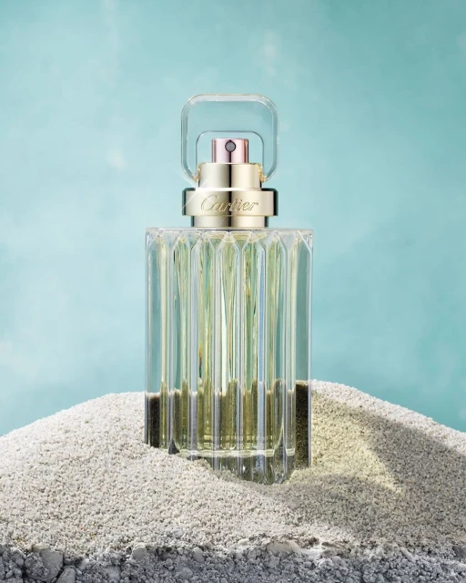 Elegant, clear glass Cartier perfume bottle centered on light gray sand against an abstract blue backdrop in a beauty still life shot.