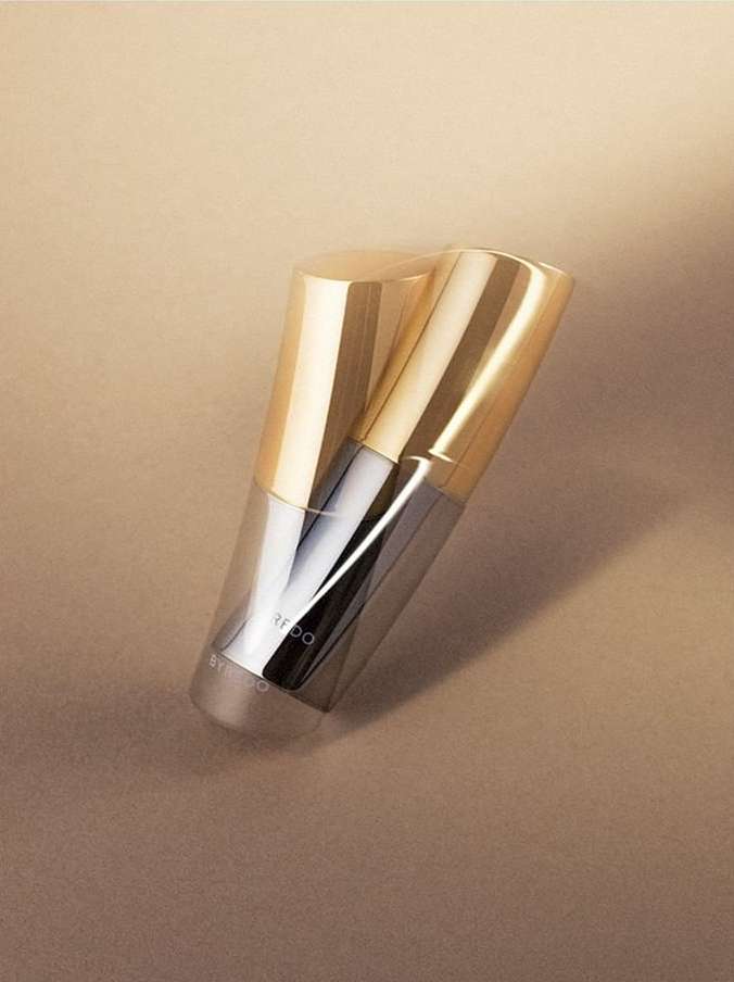 Two gold and clear cosmetic containers stand upright on a tan background in a beauty shot. The sleek product design is modern.