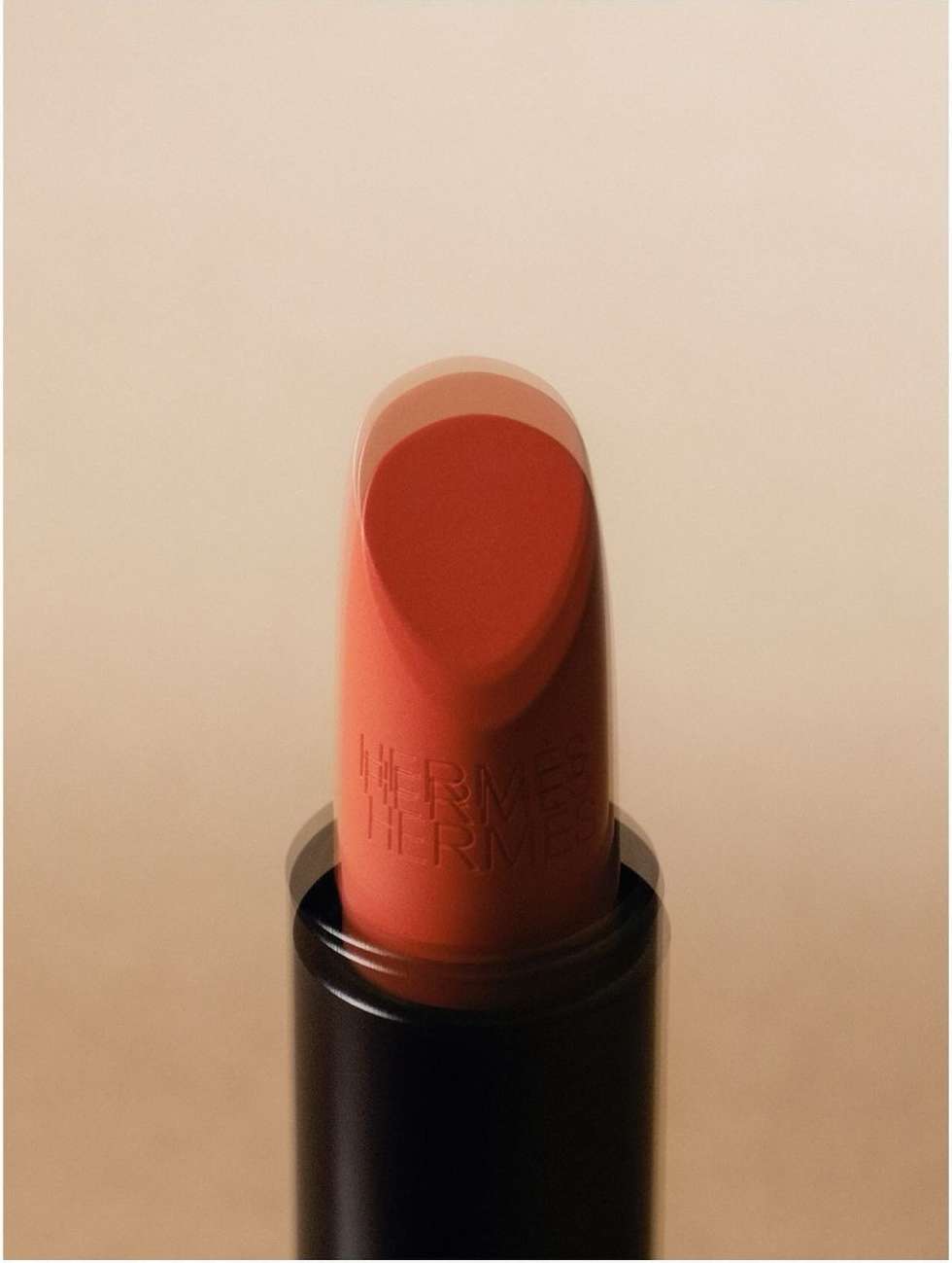 Close-up shot of a coral-red lipstick with HERMÈS engraved on it, showing the product's texture and color against a beige background.