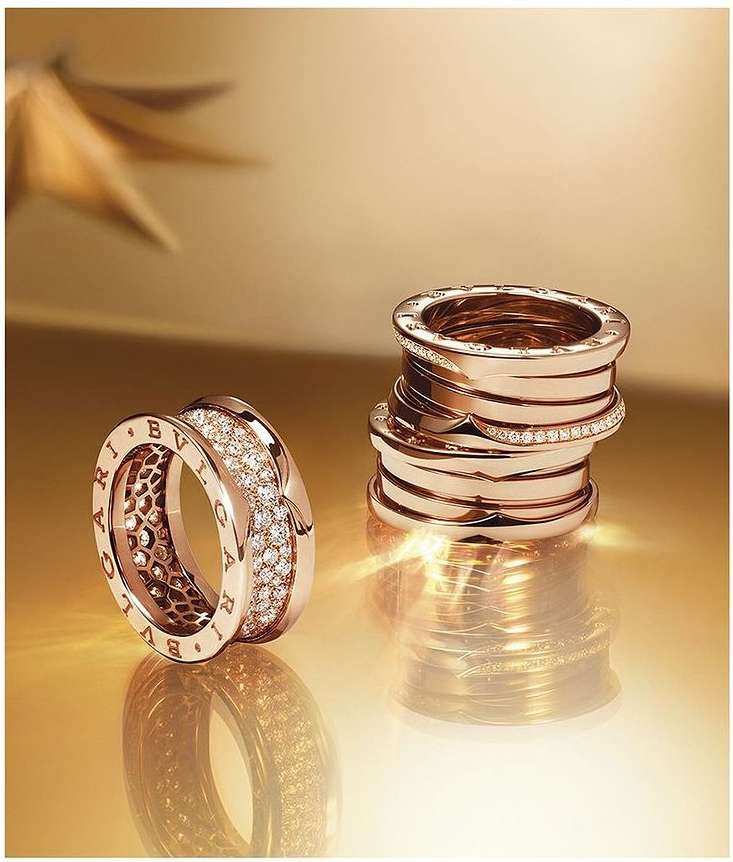 Two Bulgari B.zero1 rings on a glossy gold surface, one stacked high and one on its side showing diamond details and engraved logo.