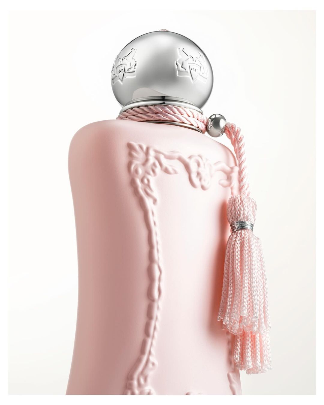 Close-up of a light pink perfume bottle with a silver cap, featuring embossed design and a decorative tassel, set against a white background.