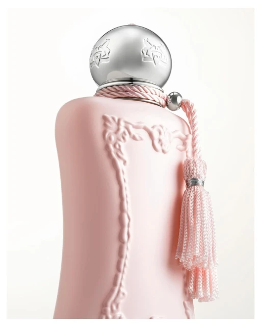Close-up of a light pink perfume bottle with a silver cap, featuring embossed design and a decorative tassel, set against a white background.