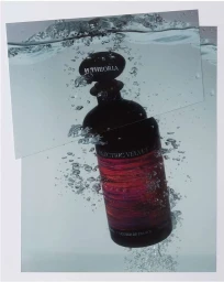 Dark glass perfume bottle submerged in water with rising air bubbles and layered photographic prints