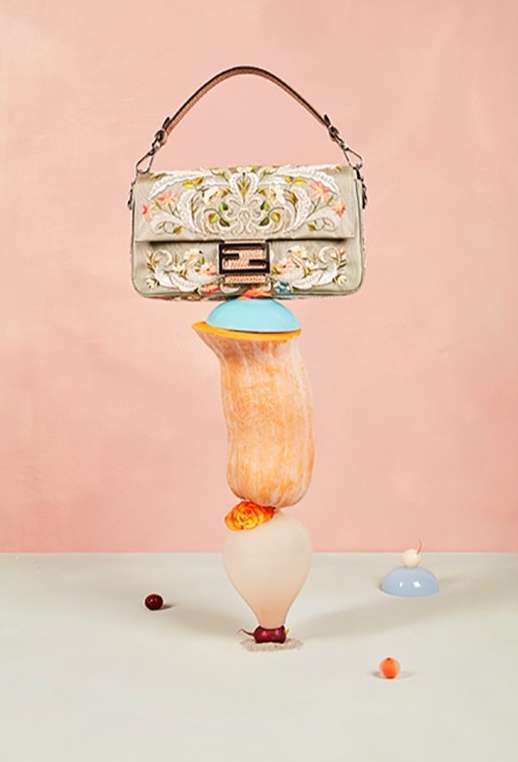 Elegant Fendi handbag balances on a tower of stylized fruit. Light pink and cream background. High-fashion product studio shot.