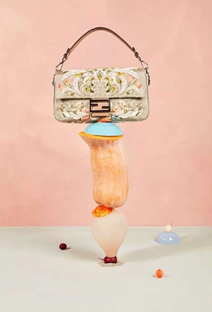 Elegant Fendi handbag balances on a tower of stylized fruit. Light pink and cream background. High-fashion product studio shot.