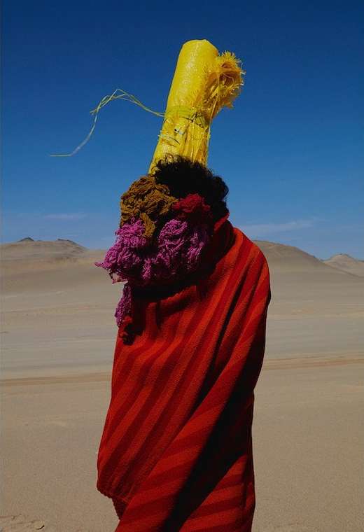 Person in a red striped poncho and ornate yellow headdress stands in the sandy desert with blue sky and distant sand dunes.