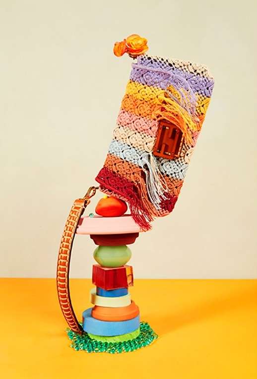 Crochet handbag with a flower accent, presented atop a colorful tower of assorted shapes against a cream and yellow background.