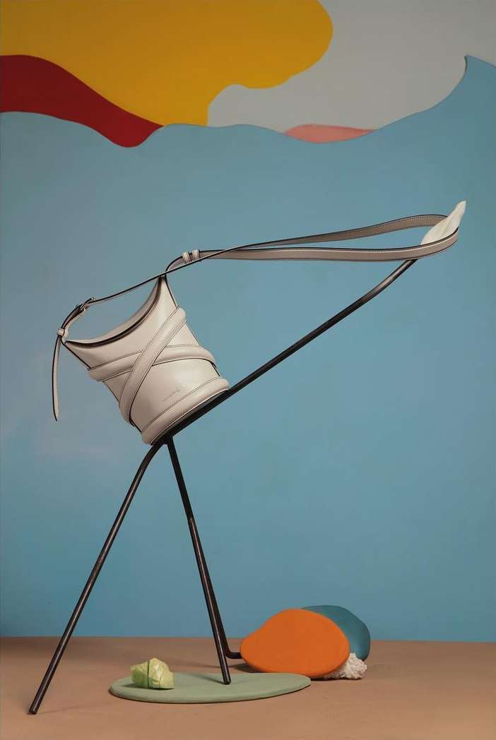 Fashion still life of a white leather purse balanced on a metal stand, with colorful abstract shapes in the background.