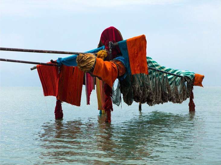 Colorful fabrics hang on a wooden structure in the sea against a bright sky, creating a striking and unusual coastal scene.
