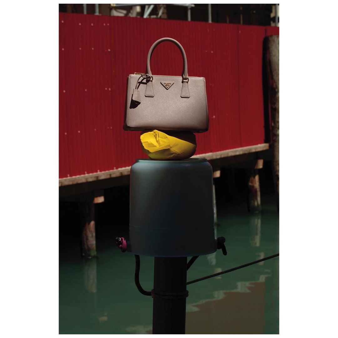 Fashion shot featuring a Prada handbag balanced on a yellow object atop a green post, set against a red wall and water.