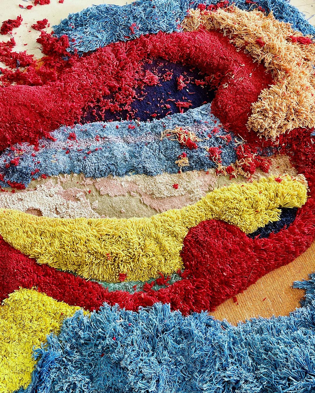 Abstract overhead view of colorful textured fiber art featuring red, yellow, and blue hues with shaggy pile and striped sections.