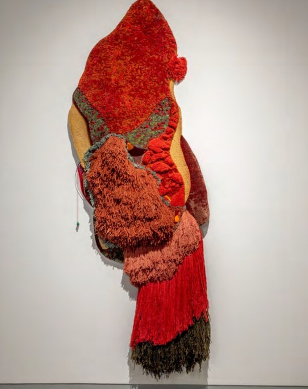 Abstract fiber art wall hanging with textures and hues of reds, brown, yellow, and green on a white wall. Interior design.