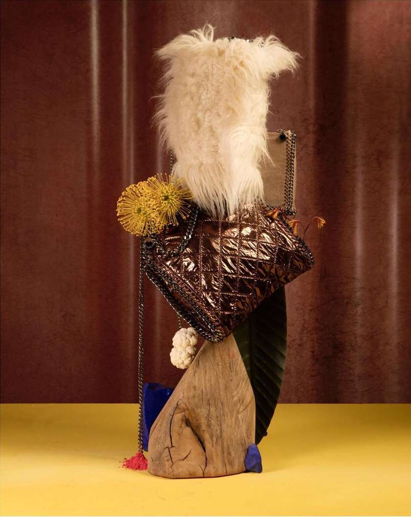 Still life shot of a brown quilted purse with a chain strap, styled with a feathery object, flowers, and wood, on a yellow surface.