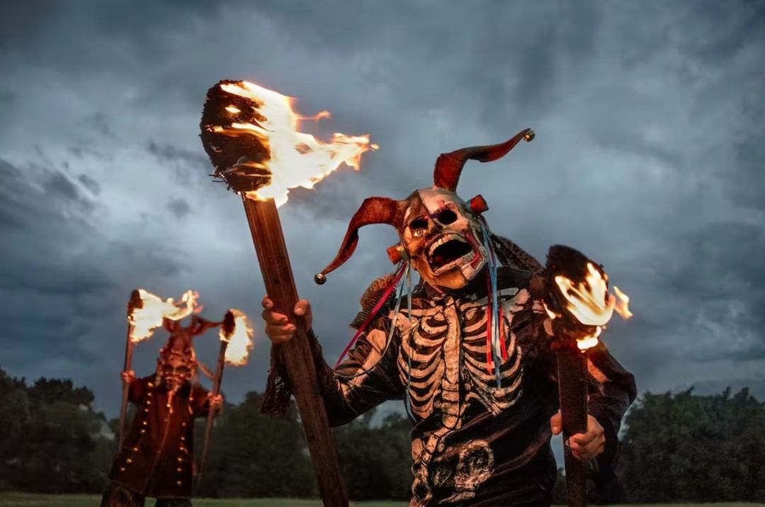 Two performers in costume with skull masks and torches pose outdoors against a cloudy sky at dusk, creating a spooky and dramatic scene.