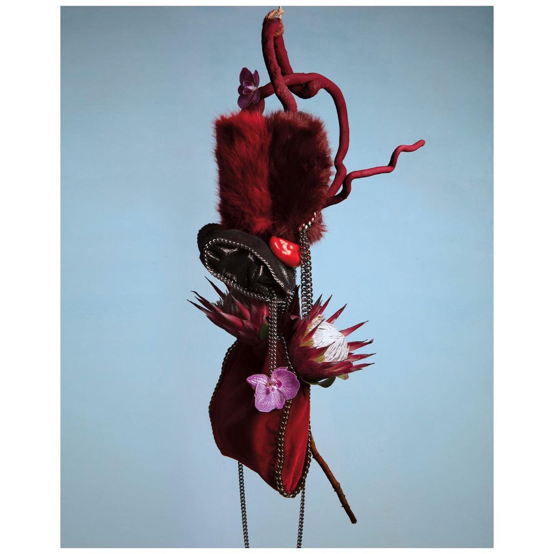 Dark red, faux fur floral arrangement with an orchid and protea, hangs in a studio setting against a solid, light blue background.