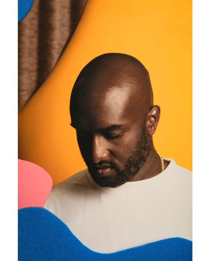 Close-up studio shot of a bald Black man in a white shirt against an orange backdrop with abstract blue and pink shapes.