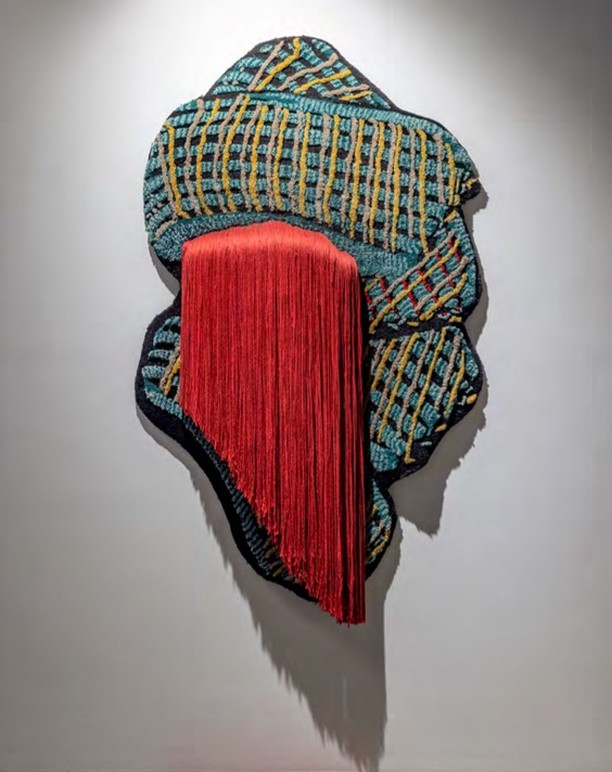 Textile art featuring black, yellow, and teal woven sections, paired with flowing vertical red fringe, on a white wall with soft lighting.