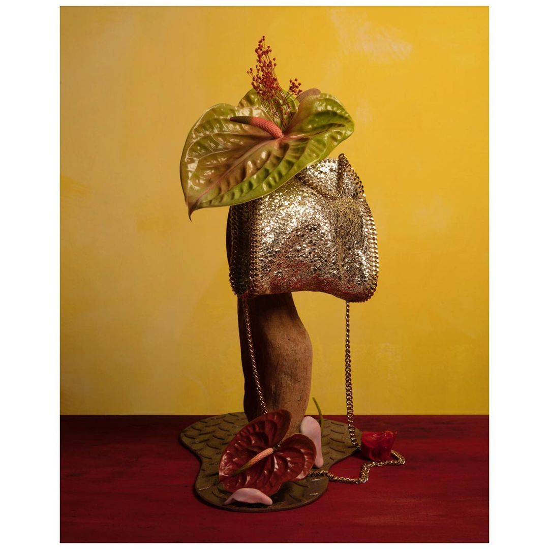 Still life photograph featuring a metallic handbag adorned with flowers, posed on a wooden stand against a golden backdrop.