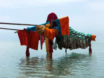 Colorful textiles and fabrics draped over a wooden framework standing in shallow water under an overcast sky