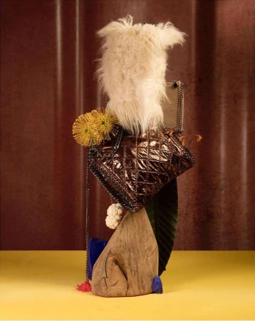 Scupltural still life assemblage featuring a quilted metallic bronze handbag, white fur, yellow flowers, and wooden elements against a textured backdrop