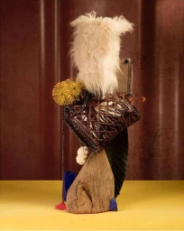 Scupltural still life assemblage featuring a quilted metallic bronze handbag, white fur, yellow flowers, and wooden elements against a textured backdrop