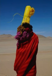 Figure draped in a red blanket wearing a tall yellow cylinder and colorful yarn mask in a desert