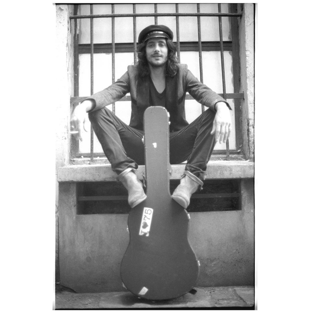 Black and white portrait of a long-haired man with a cigarette seated on a ledge with a guitar case in front of a barred window.