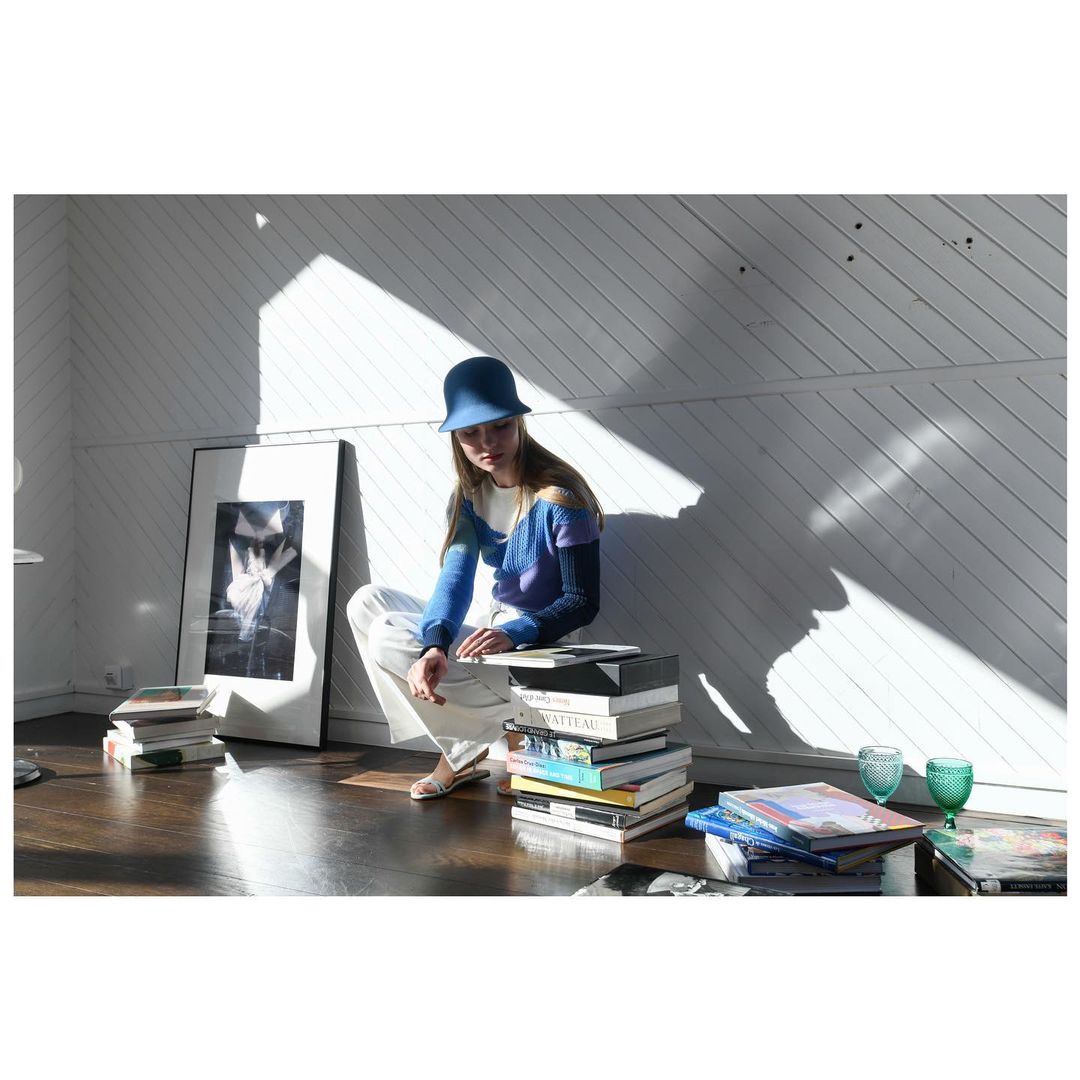 Woman in a blue hat sits on a stack of books in a bright room with diagonal shadows and a black and white framed print.