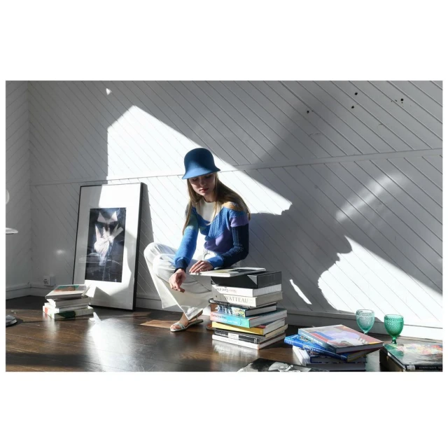 Woman in a blue hat sits on a stack of books in a bright room with diagonal shadows and a black and white framed print.