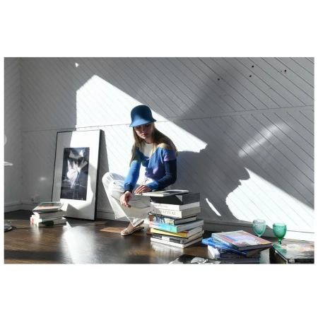 Woman in a blue hat sits on a stack of books in a bright room with diagonal shadows and a black and white framed print.
