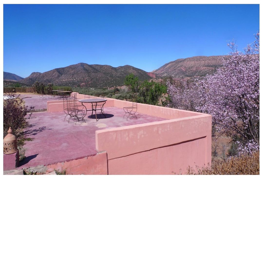 Outdoor terrace with metal table and chairs overlooks a valley with mountains and flowering trees under a clear blue sky on a sunny day.