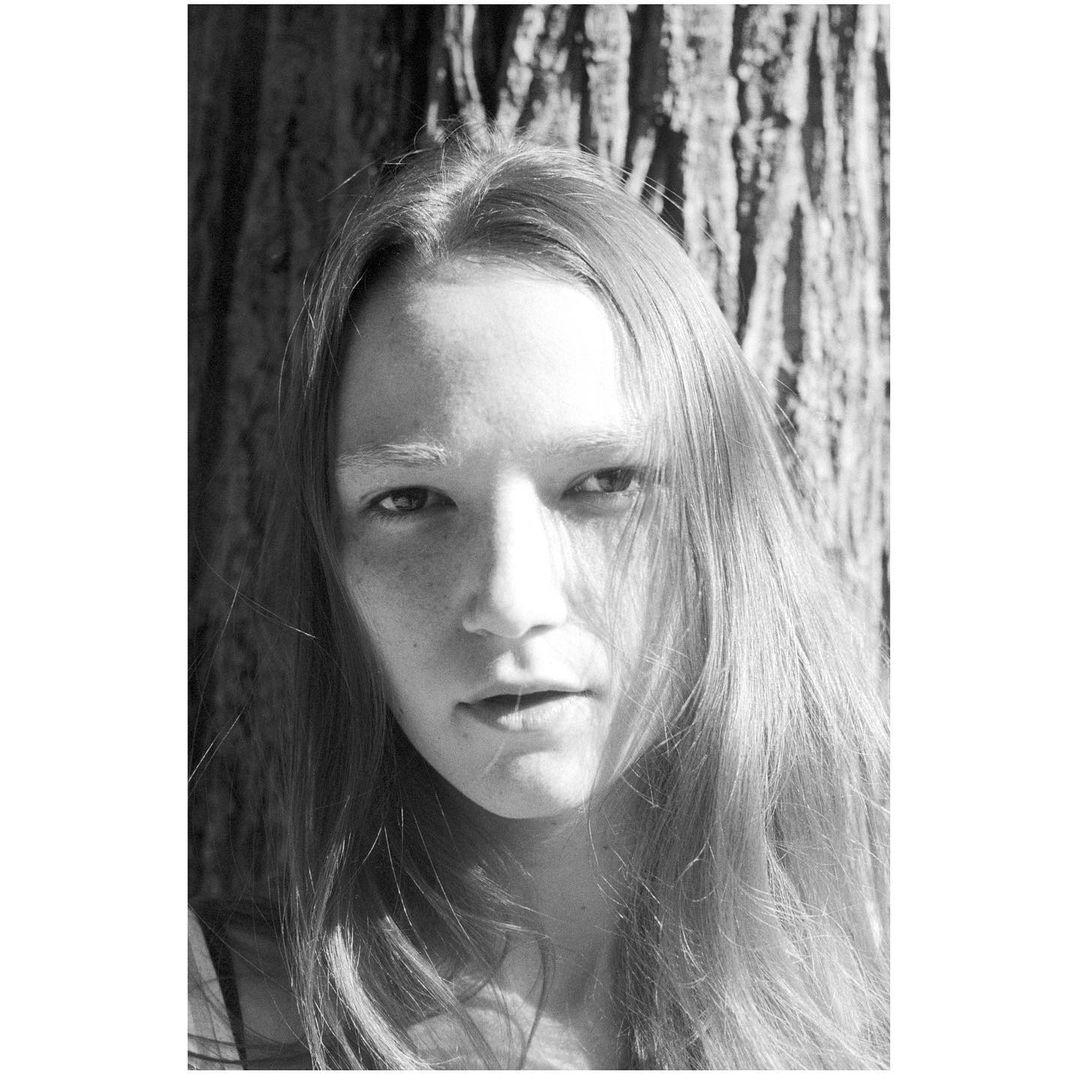 Black and white portrait of a young woman with long hair, facing the camera, with a blurred tree trunk visible in the background.