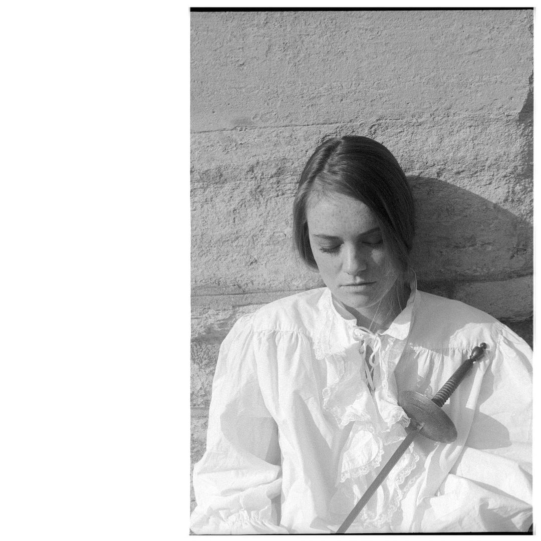 Monochrome portrait of a young woman with a fair complexion in a white ruffled shirt holding a fencing foil, stone wall background.