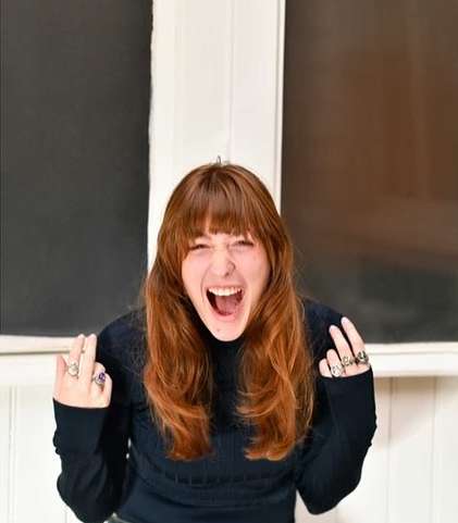 Close-up of a young, light-skinned woman with auburn hair, wearing black long-sleeve shirt, and multiple rings, shouting with hands raised indoors.