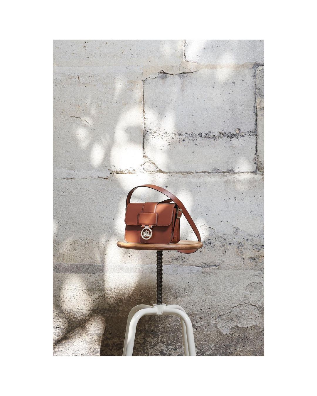 Brown leather purse with silver hardware sits on a white industrial stool against a textured grey wall in an indoor setting.