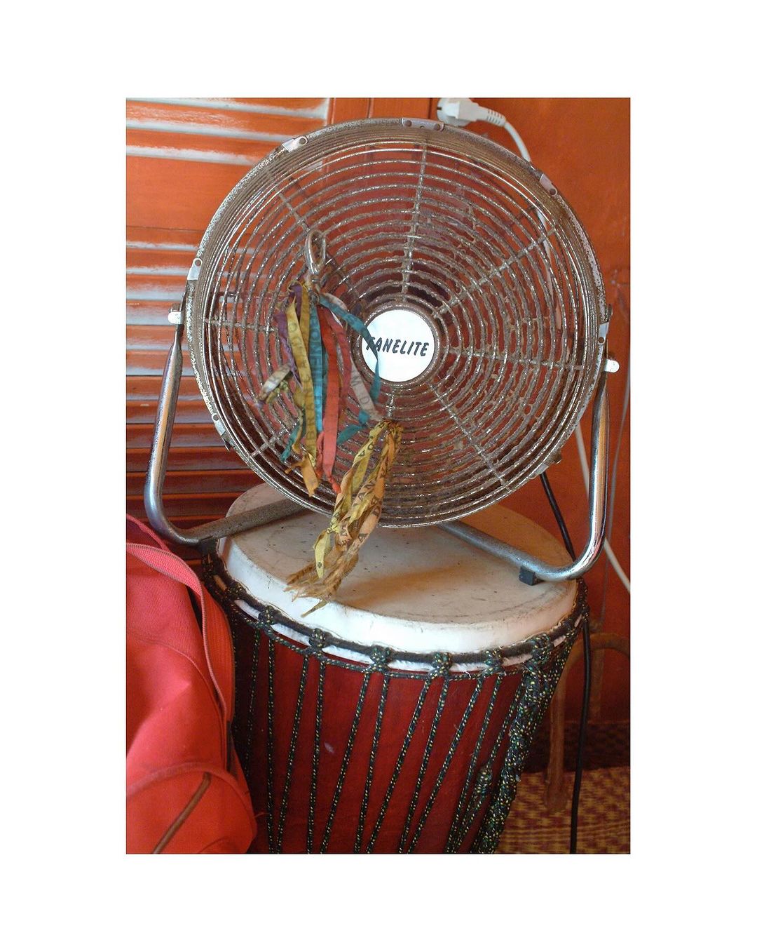 Chrome-finish fan sitting on a drum with hanging ribbons, against an orange wall, in an indoor setting with soft lighting.